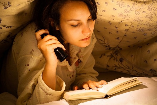 Serious Woman Reading a Book with Flashlight on Bed